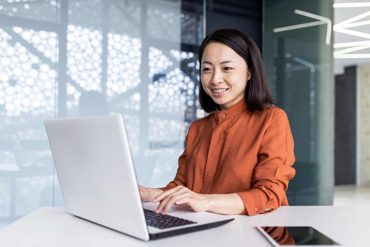 Successful asian business woman working inside office building with laptop, female worker smiling and looking at computer screen, satisfied with work achievement results